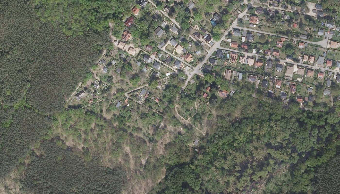 Luftbild einer Ortslage am Waldrand: Einfamilienhäuser mit Gärten, daneben unbebaute Flächen und geschlossener Kiefernwald.